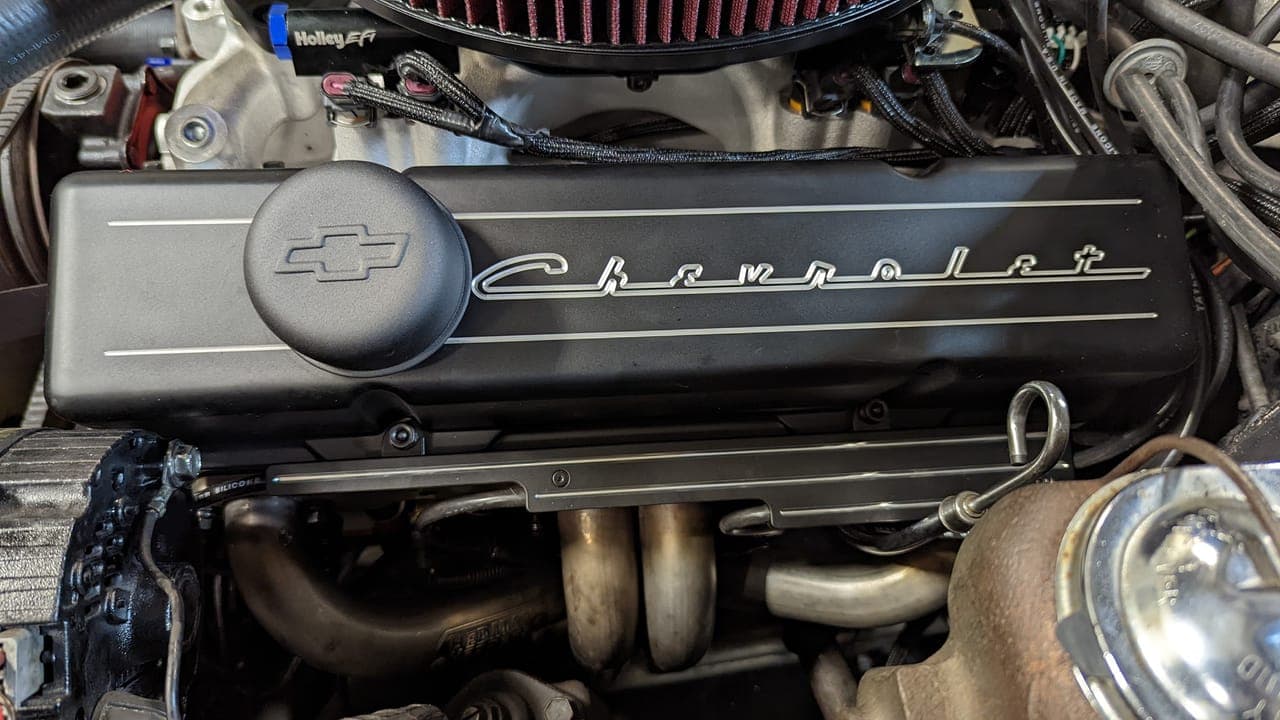 Close-up of a black small-block Chevy valve cover with the silver Chevrolet script and bowtie logo, headers visible below