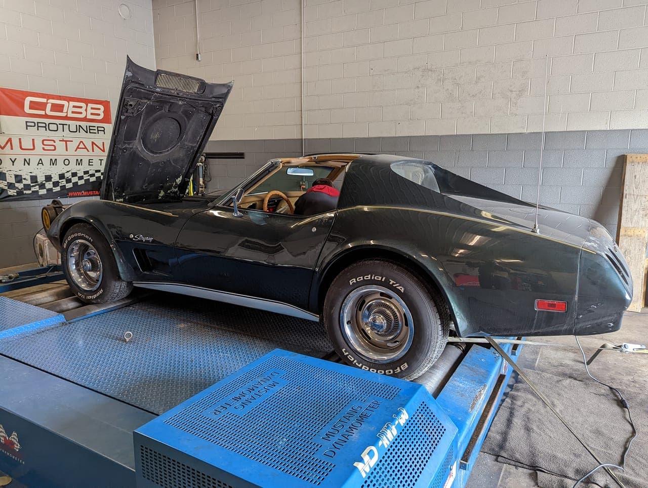 Dark green 1974 Corvette strapped down on a Mustang chassis dynamometer with its hood up inside the dyno bay