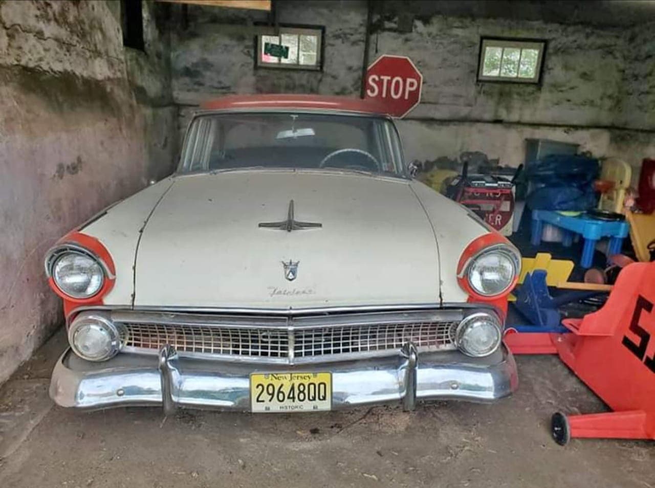 Original red-and-white 1955 Ford Fairlane sitting dusty inside a stone garage as a barn find, not running