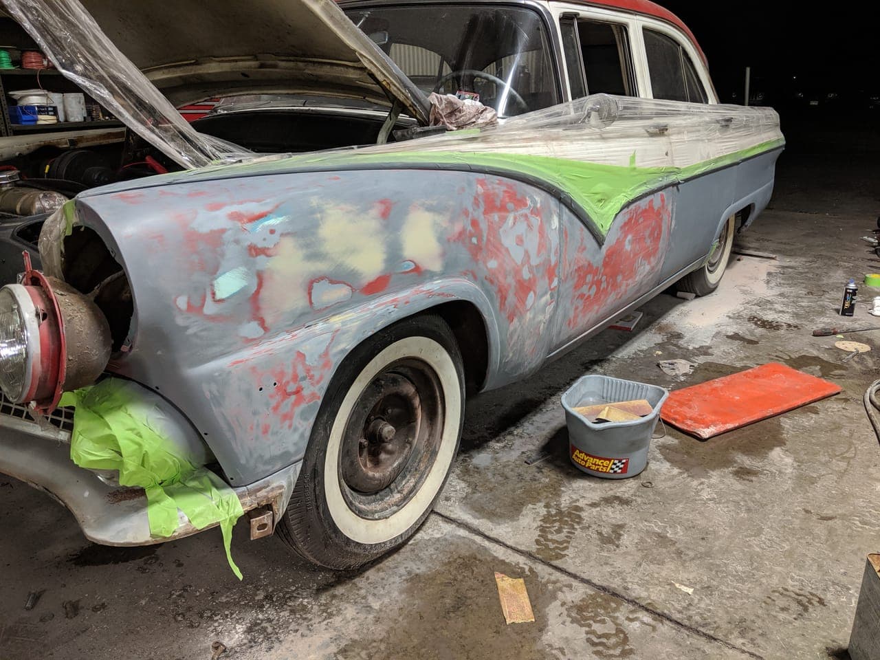 1955 Ford Fairlane stripped to gray primer in the shop at night, hood up and panels sanded with the original red still showing through
