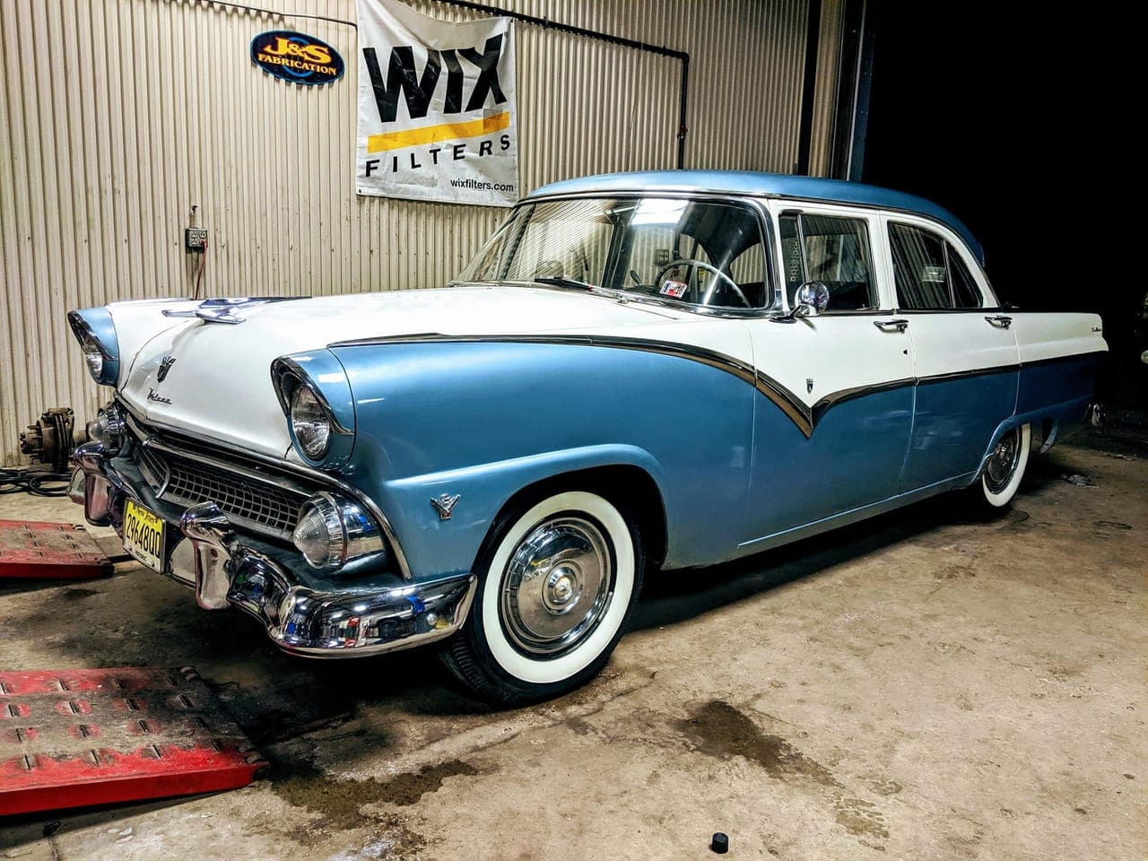 Finished blue-and-white 1955 Ford Fairlane with whitewall tires and polished chrome on the shop rack under dramatic lighting