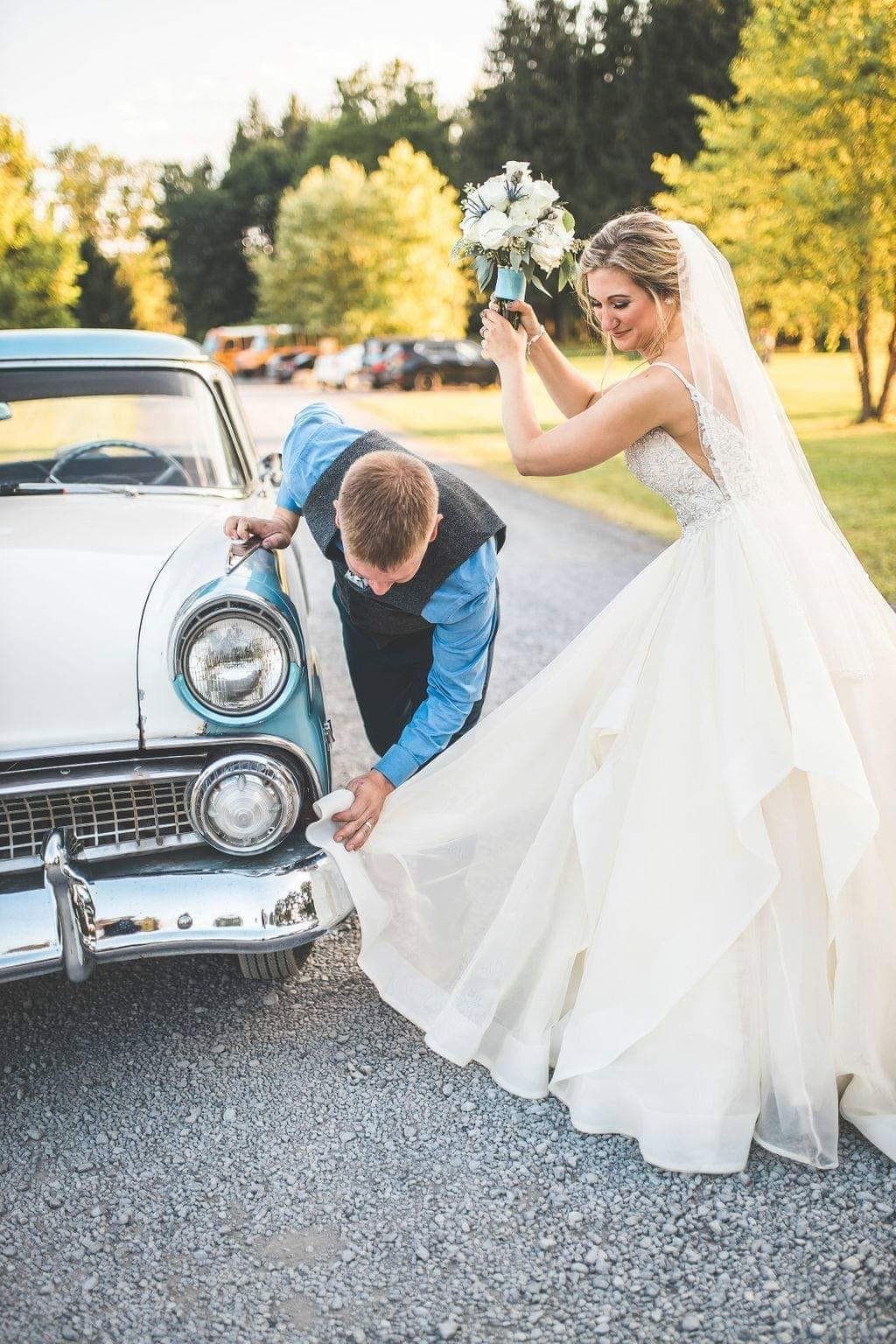 Groom in a vest crouched at the front fender of the blue 1955 Fairlane while the bride in her gown holds up her bouquet