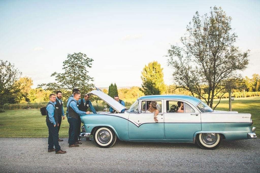 Blue-and-white 1955 Ford Fairlane on a wedding day, groomsmen gathered at the open hood and the bride leaning out the window at golden hour