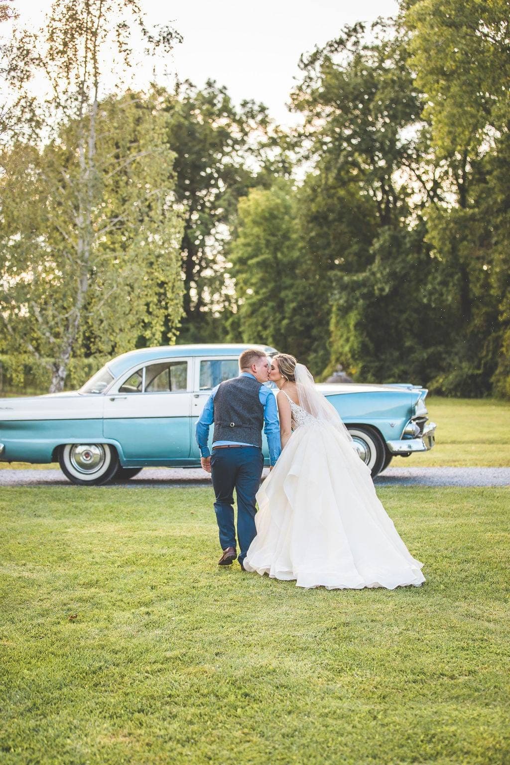 Bride and groom kissing on a lawn in front of the two-tone blue 1955 Ford Fairlane at golden hour