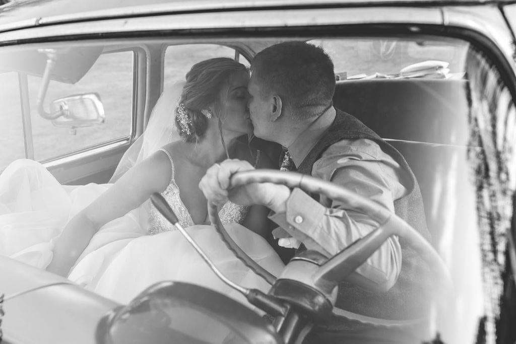 Black-and-white photo of the bride and groom kissing behind the wheel of the 1955 Ford Fairlane