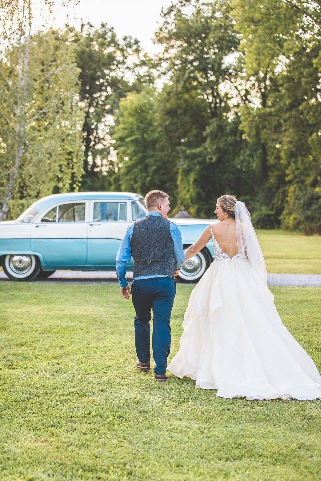 Bride in her gown and groom in a vest walking hand in hand across a lawn toward the blue 1955 Ford Fairlane