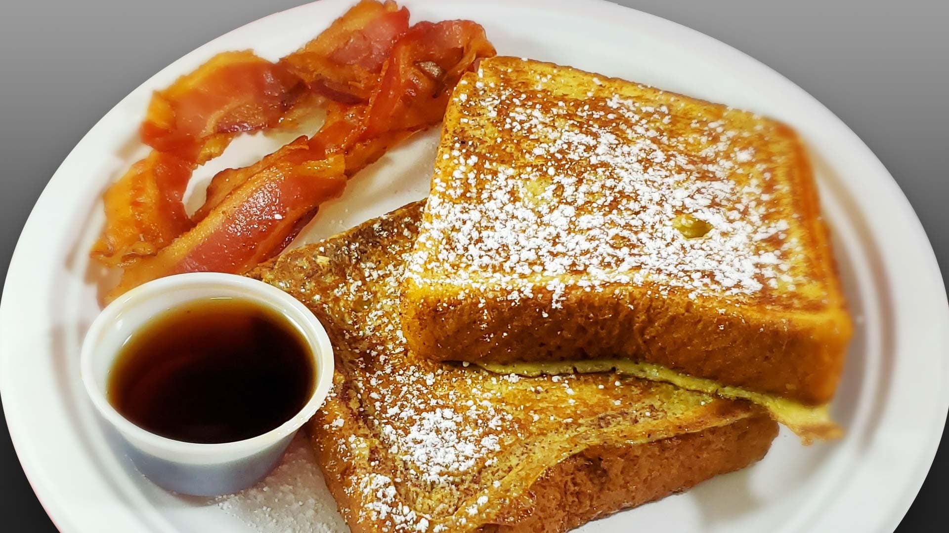 Powdered-sugar French toast with a side of bacon and a cup of syrup at Cheezie Sam's Grill