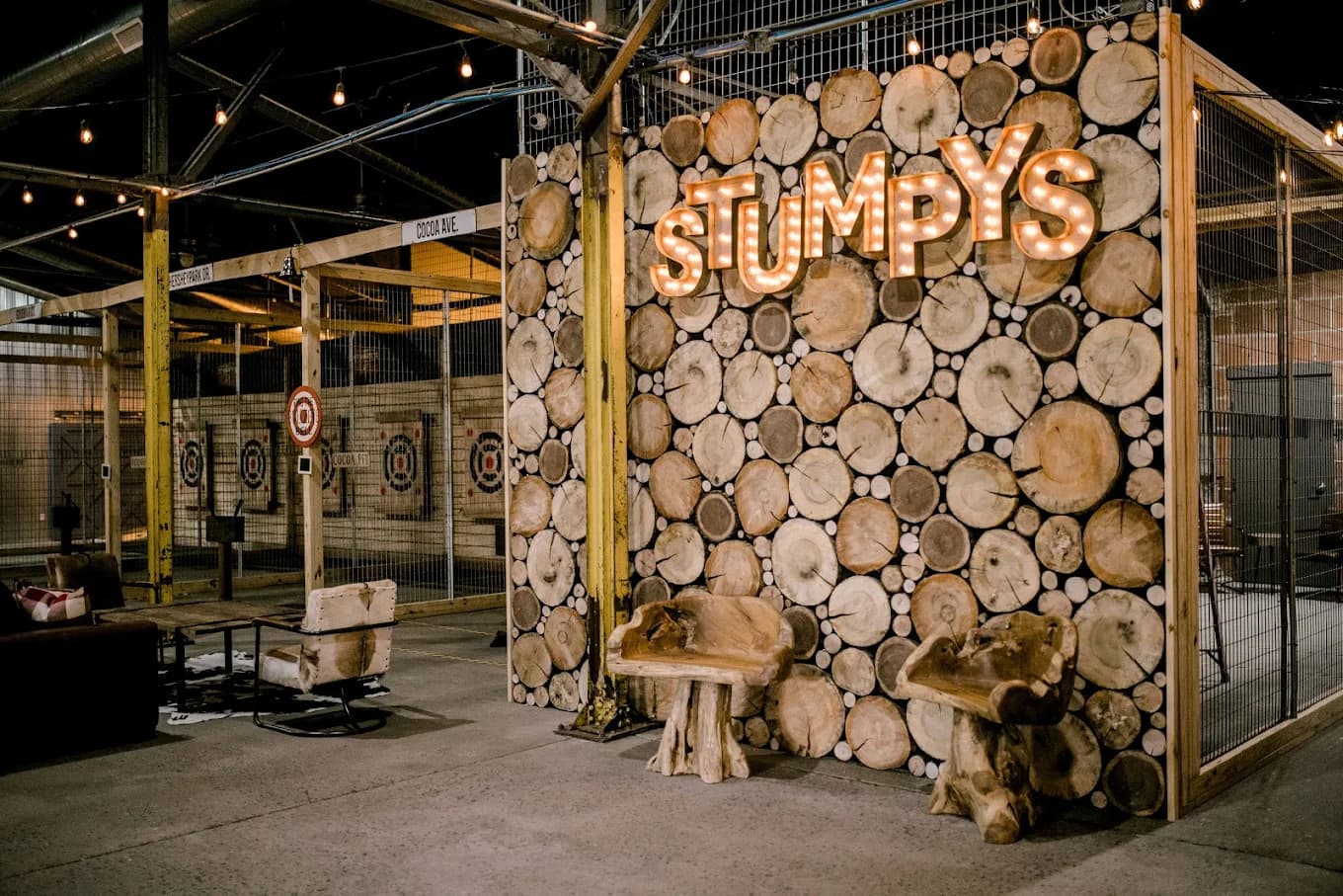 Caged axe-throwing lanes with bullseye targets and an illuminated 'STUMPYS' sign on a wall of stacked wood rounds at Stumpy's Hatchet House of Hershey