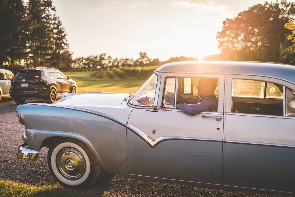 Groom in a vest driving the finished two-tone Stratocaster-blue 1955 Ford Fairlane down a country lane at golden hour, the sun flaring behind the car
