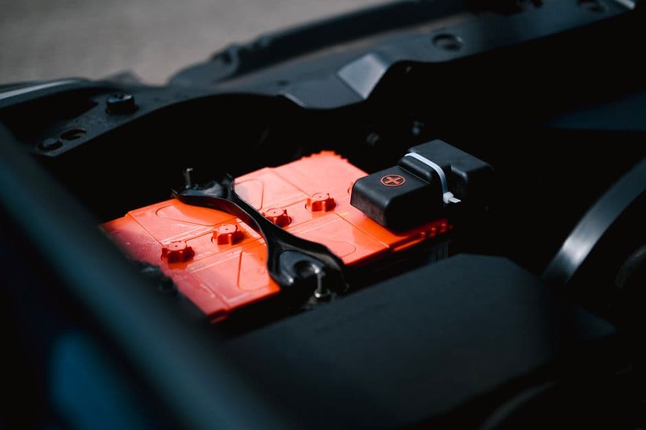 Detailed view of an orange car battery inside a vehicle engine bay during hot-weather battery testing.